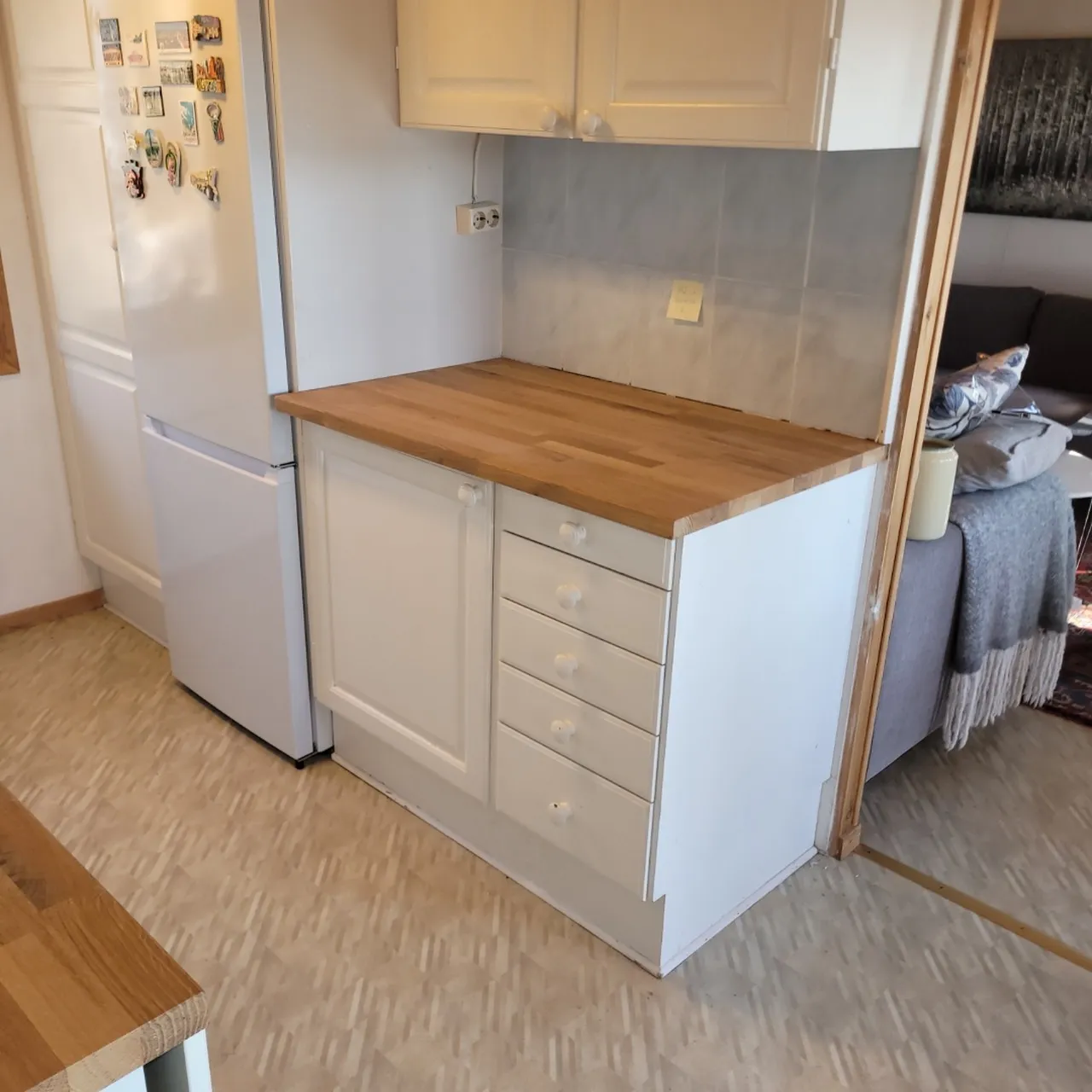 Ett litet kökshörn med vita skåp, en bänkskiva i trä, fyra lådor, ett kylskåp med magneter och ett kaklat stänkskydd; ett vardagsrum med en grå soffa syns i bakgrunden.